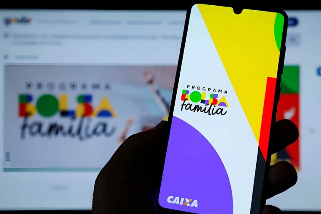 Uma mão segurando um celular com o aplicativo do Bolsa Família aberto. Ao fundo, a tela de um computador com o site para verificar aumento do Bolsa Família.