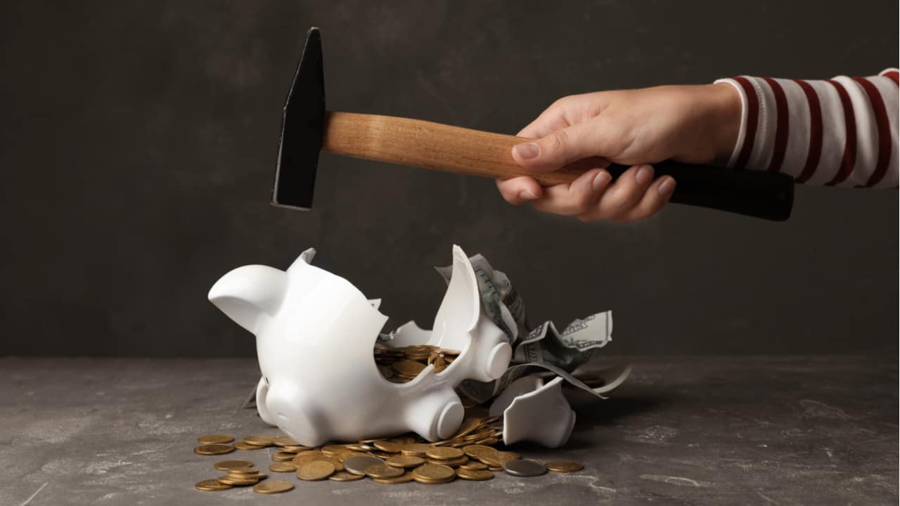 Pessoa quebrando cofre de poupança com martelo e moedas espalhadas